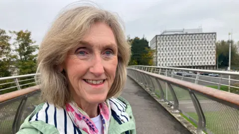 Maureen Millar devant un pont menant à Marlborough House, coiffée d'un manteau vert et d'une écharpe à motifs roses.