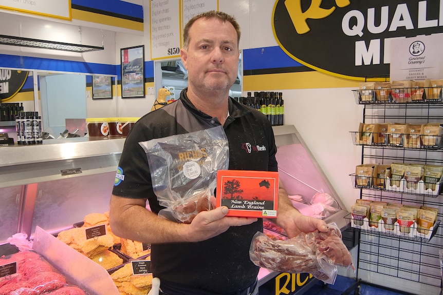 Un homme en t-shirt noir montrant différentes coupes de viande devant un présentoir réfrigéré.