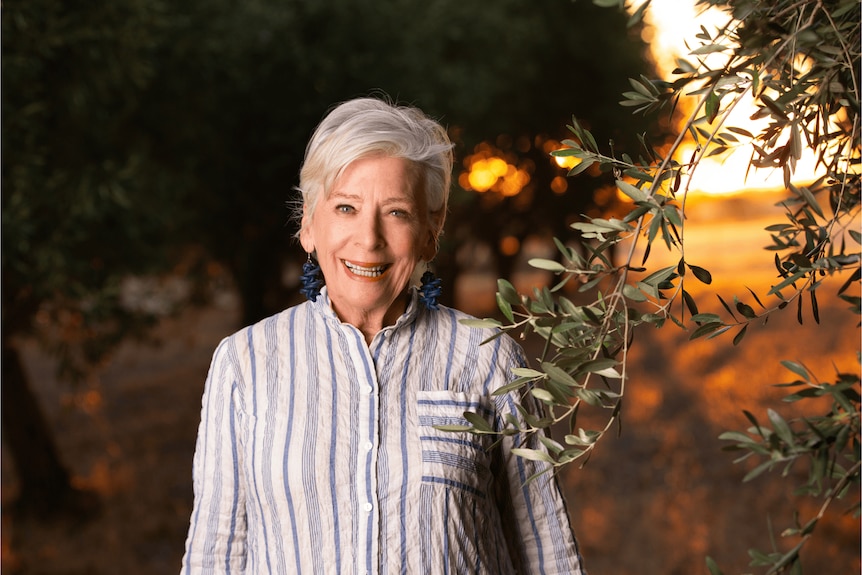 Maggie Beer souriante devant des arbres, avec un coucher de soleil derrière elle.