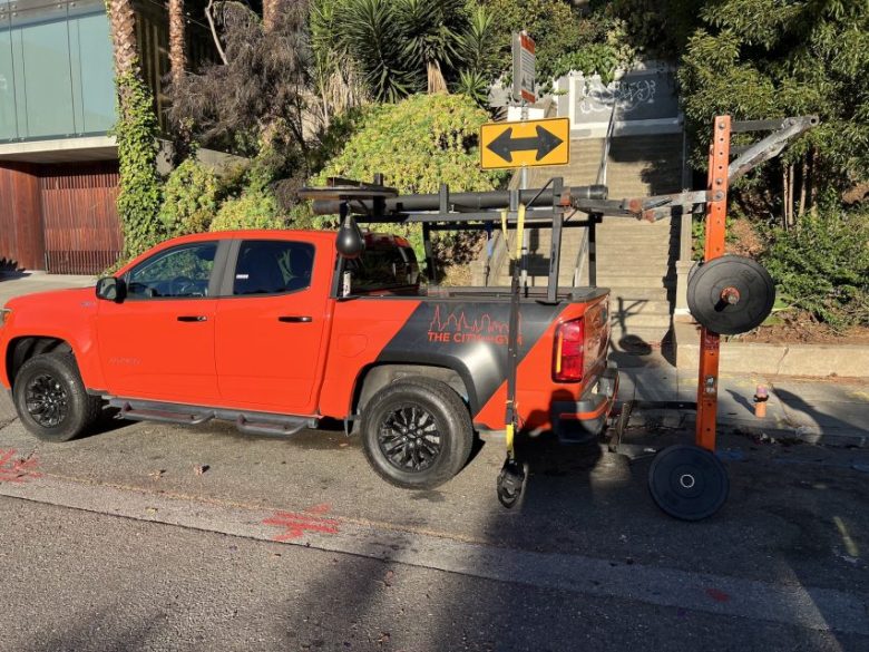 Un camion rouge avec un équipement de gym dans son bed, garé sur une rue près des escaliers et de la verdure.