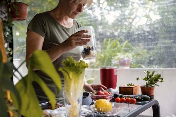 Une femme tient un récipient avec des baies et des tranches de banane ; d'autres fruits et légumes sont disposés sur une table devant elle.