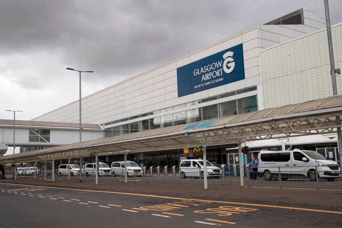 Vue extérieure d'un terminal d'aéroport sous un ciel nuageux, avec plusieurs véhicules garés devant le bâtiment, un grand panneau bleu visible sur le côté du terminal.